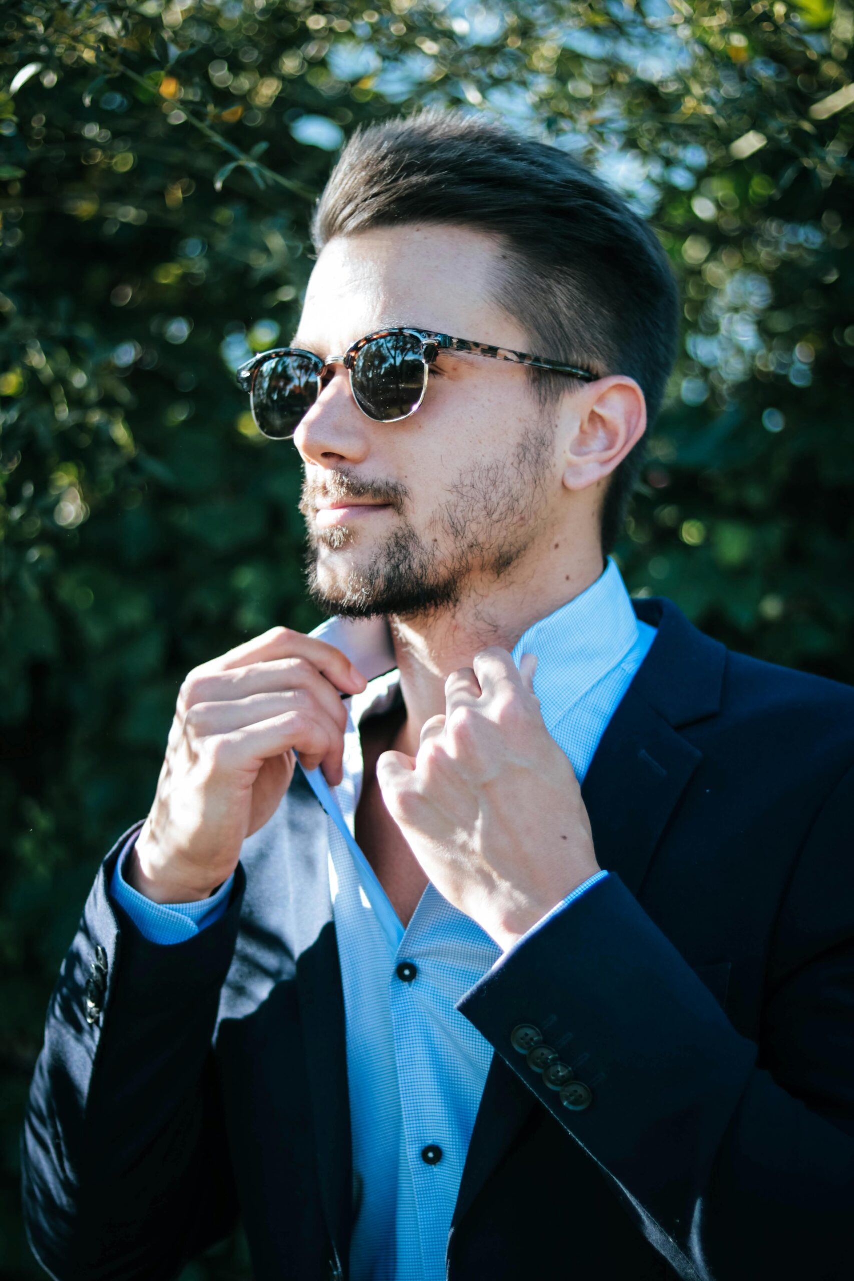 Fashionable man adjusting his suit and sunglasses outdoors on a sunny day.