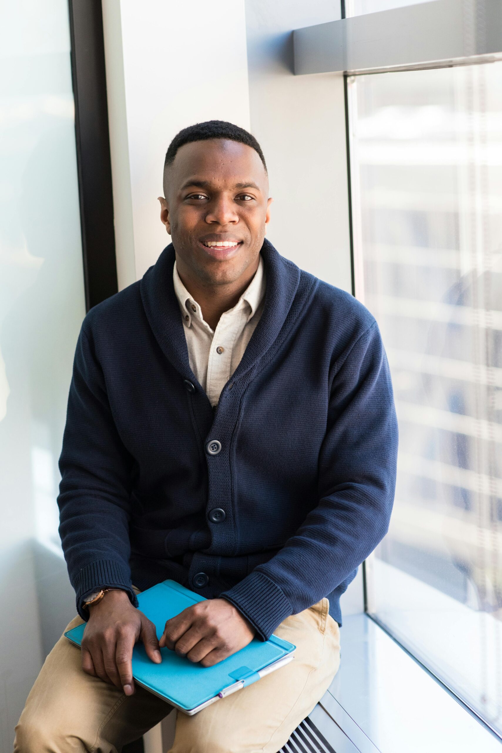 A professional black man smiling while seated by a window, embodying modern business vibes.