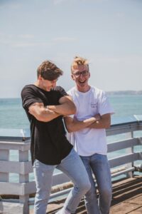 Two men laughing and posing outdoors against a sea backdrop, exuding stylish vibes.