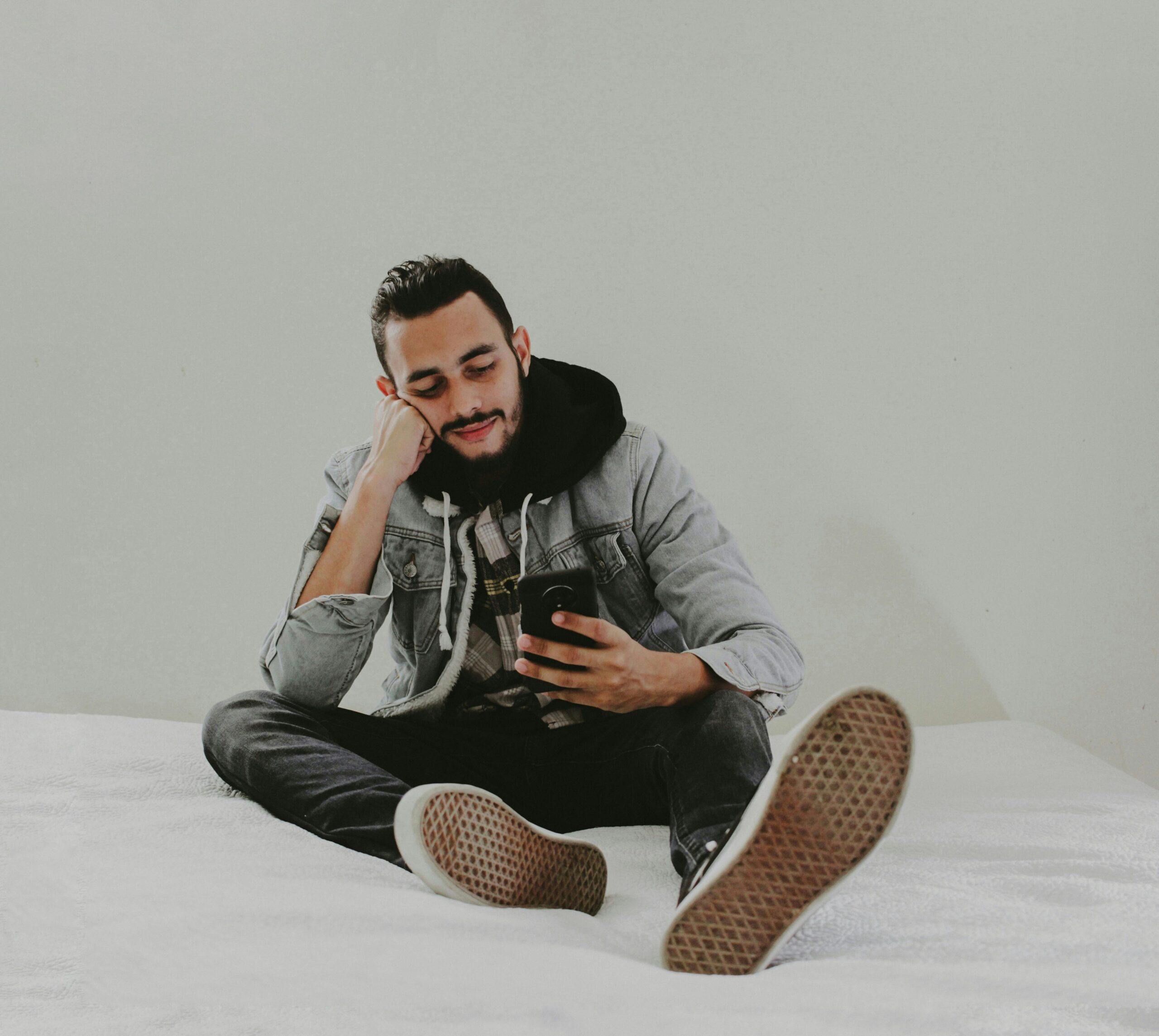 Man in casual wear sitting on bed with smartphone, wearing hoodie and denim jacket.