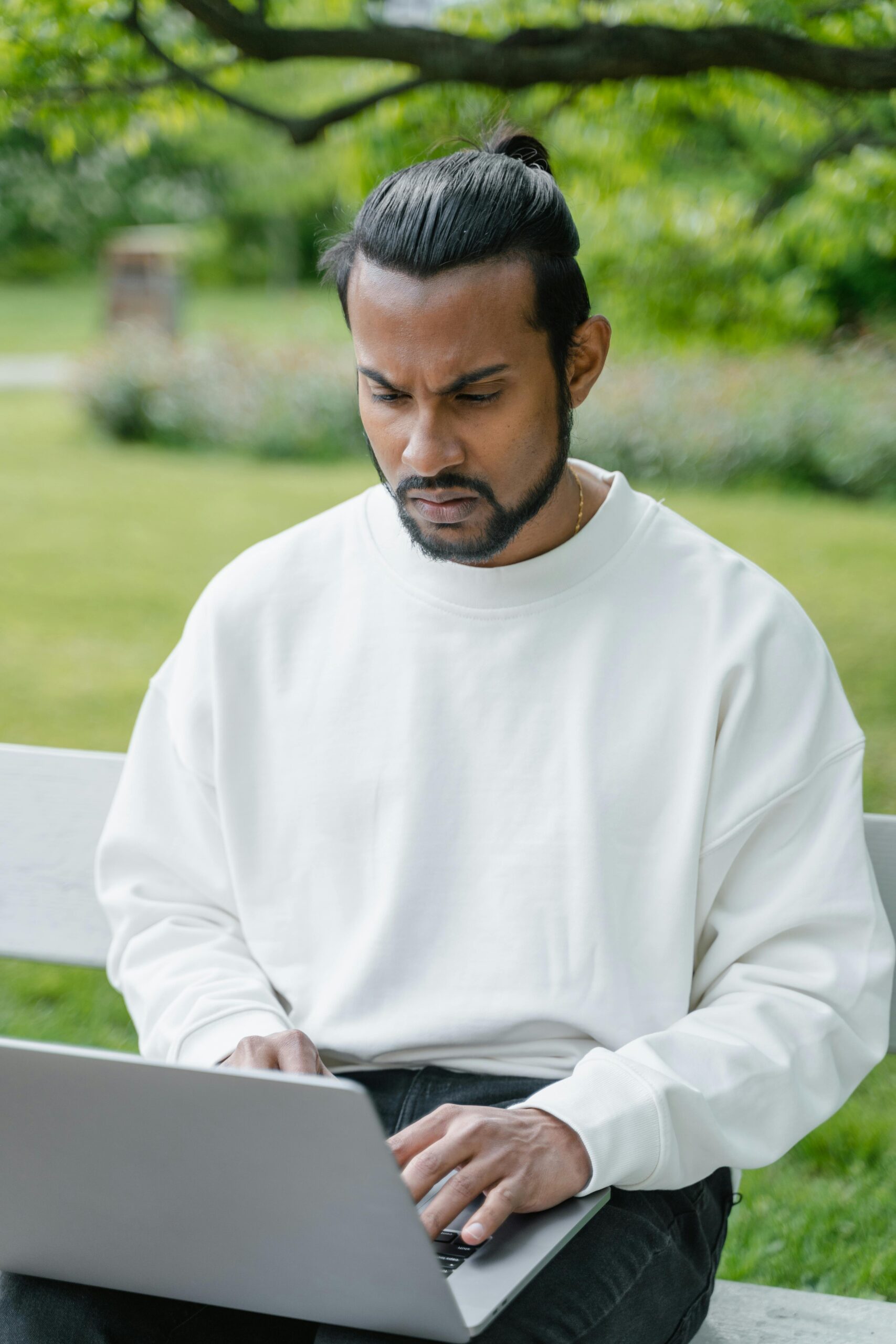 A focused young man in a white sweater works on his laptop outdoors in a park.